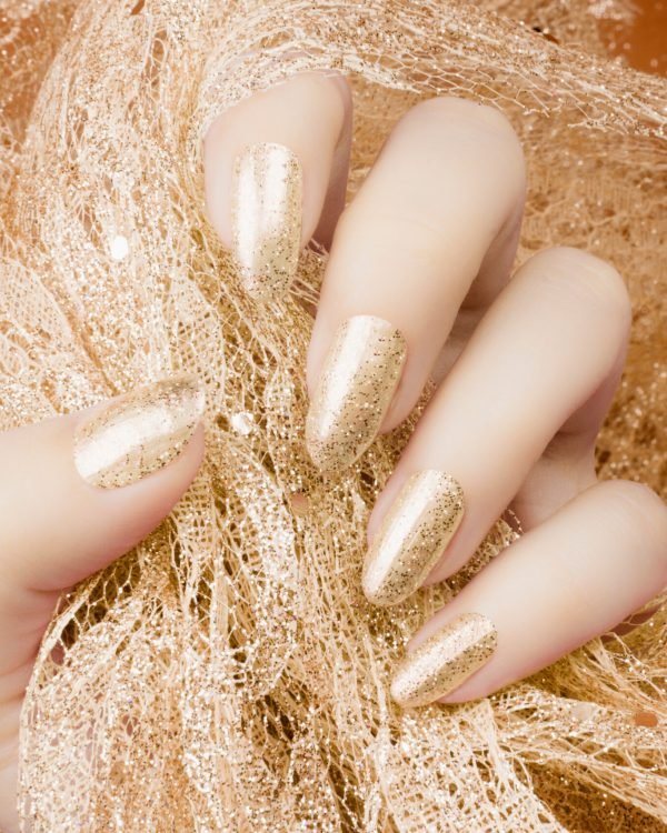 Female hand with glittered gold nails manicure is holding gold decoration on orange background.